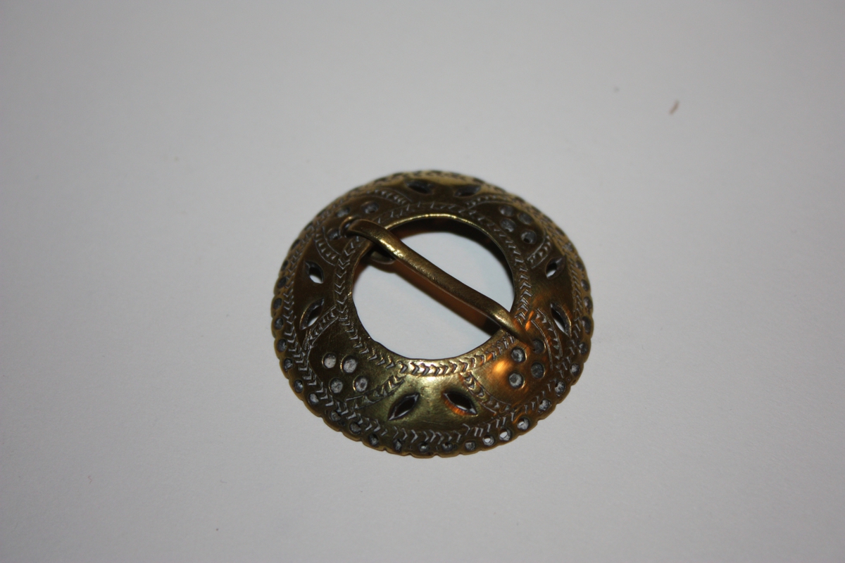 Ringforma med gjennombroten dekor og bølgja kant med hol innanfor. Gravert. Torn på oversida. Nytta som skjorteknapp