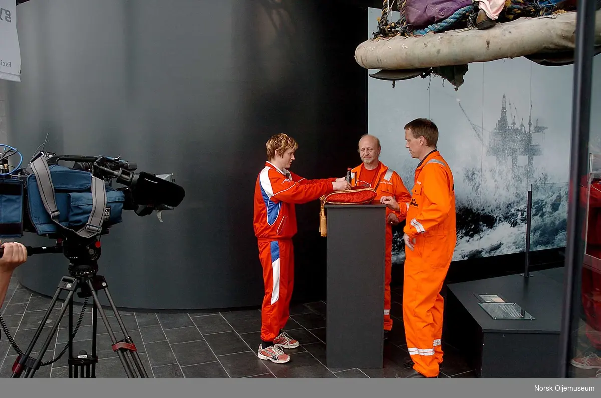 Flaske med "Den første olje" -  råolje fra det som skulle bli Ekofiskfeltet, skal stilles ut på Norsk Oljemuseum i Stavanger.
Anledningen er TV-opptak til NRK-programmet "Norsk Attraksjon" som ble vist på TV i september 2009.       
Programskaper Linda Eide t.v. danderer flasken med råolje sammen med gjenstandsansvarlig Svein Terje P. Førland samt museumsdirektør Finn E. Krogh t.h. i bildet.