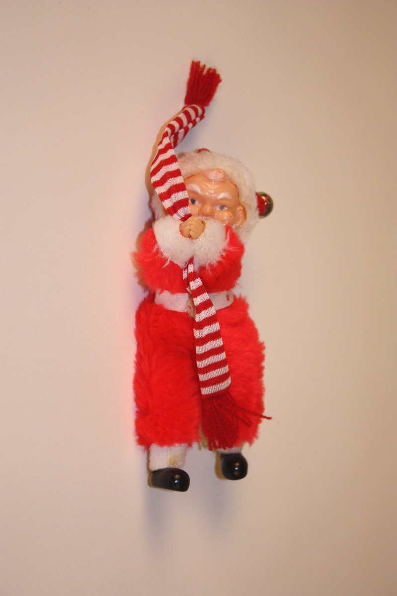 Nisse i heil flossdrakt. Kvitt belte og kantingar. Ansikt av plast. Bjelle i toppen av hua. Nissen held på eit langt sjal i raude og kvite striper.