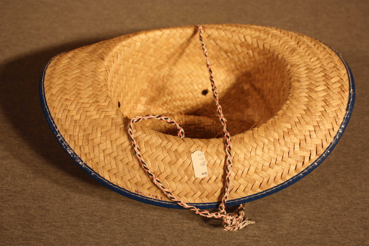 Stråhatt med påsydd blå plastlist rundt bremmen og plaststjerne og plasthestesko festa på pullen. Bomullstau som hakeband