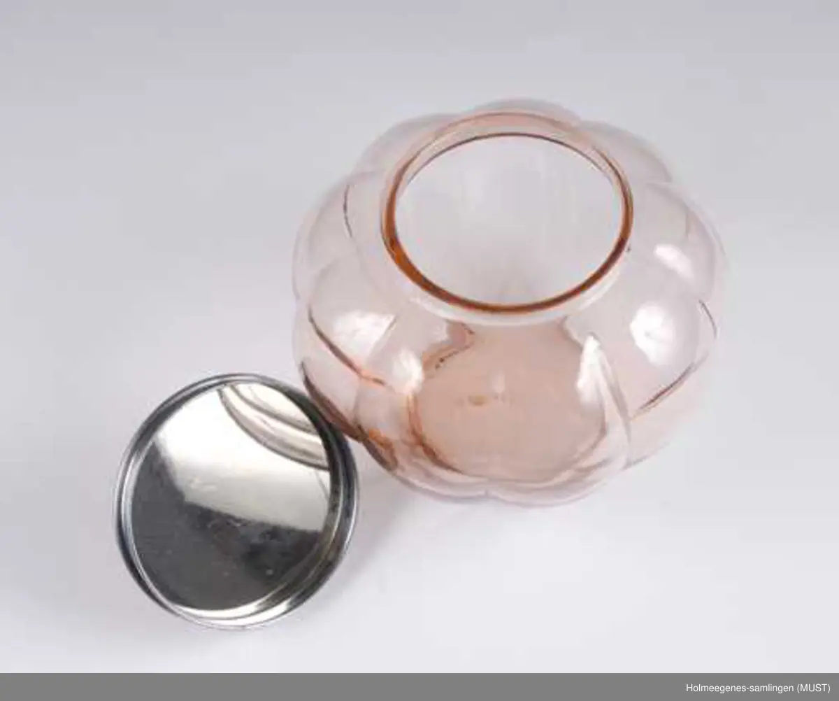 Bolle av formpresset rosa glass. Lokk av blikk med guillochering, belagt med rosa plast med knott av plastlignende materiale. (Guillochering er en maskinell metode å behandle sølv eller lignende materiale for å gi den et regelmessig rillemønster eller et geometrisk mønster som skimrer gjennom emaljen, her plasten.