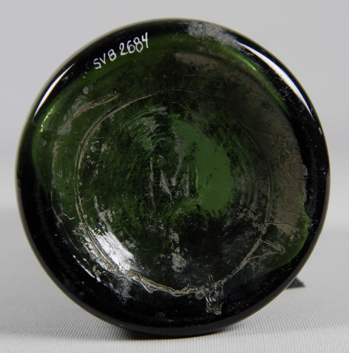 Drikkeflaske i grønt, klart glass. Merket "M" på flaskebunnen. En del leire på flasken. 