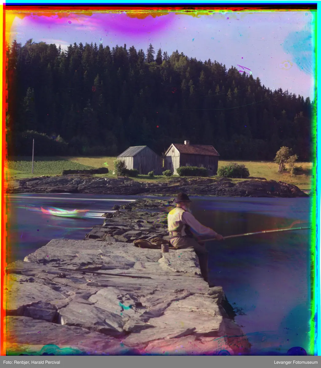 En mann sitter og fisker på Jeteen. I bakgrunnen sees husmannsplassen "Nesset", Levanger.