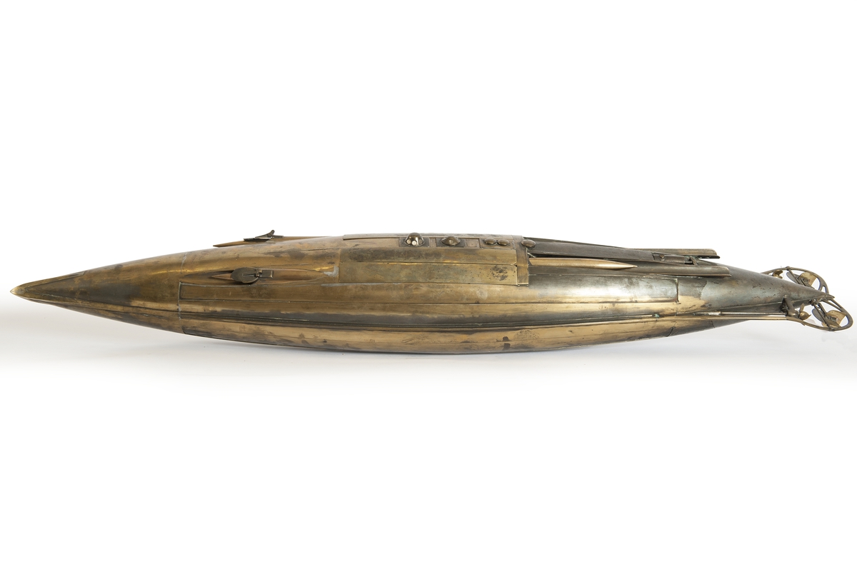 Spolformad. Stålplåt. Slingerköl, 2 propellrar av metall, 2 st höjd- och sidoroder parallellkopplade med gardjärn. 2 st höj- och sänkbara periskop av mässing. 4 st rännor för torpeder, därav 2 med luckor framtill, dels 2 för magasinering. Kort cylinder, rörlig i ränna midskepps akterut. Torpedrännorna täckas av långa stållock. Tillhörande 4 st torpeder av massiv mässing. Eneroths förslag till ubåt 1897. UndervattenstorpedFöremålets form: Spolformad.