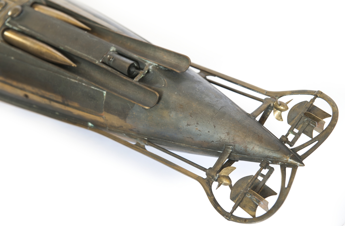 Spolformad. Stålplåt. Slingerköl, 2 propellrar av metall, 2 st höjd- och sidoroder parallellkopplade med gardjärn. 2 st höj- och sänkbara periskop av mässing. 4 st rännor för torpeder, därav 2 med luckor framtill, dels 2 för magasinering. Kort cylinder, rörlig i ränna midskepps akterut. Torpedrännorna täckas av långa stållock. Tillhörande 4 st torpeder av massiv mässing. Eneroths förslag till ubåt 1897. UndervattenstorpedFöremålets form: Spolformad.