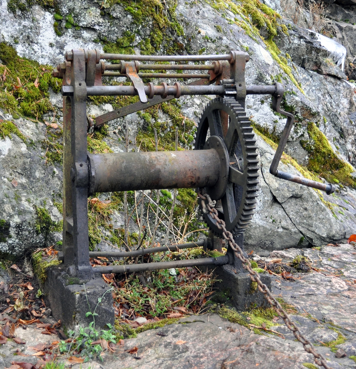 Håndvinsj av jern. Med tannhjul, sveiv og kjetting. Står på et støpt fundament.