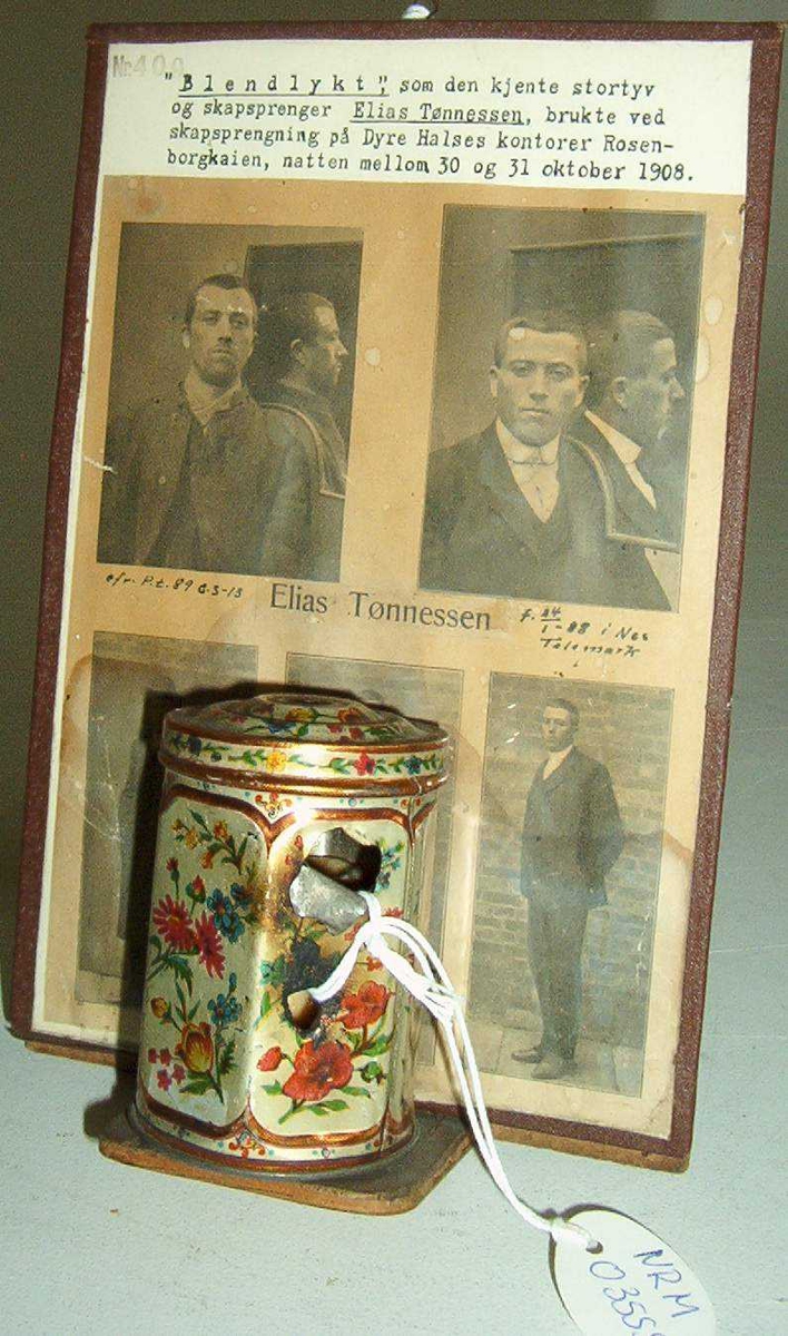 Montasje med 5 foto av Elias Tønnesen på en plate. Platen med bilder og tekst har nederst en påfestet plate for utstilling av "lykten", som er en sekskantet teboks, dekorert med blomster, og med to små hull for lyset. Teboksen inneholder en talglysstump.