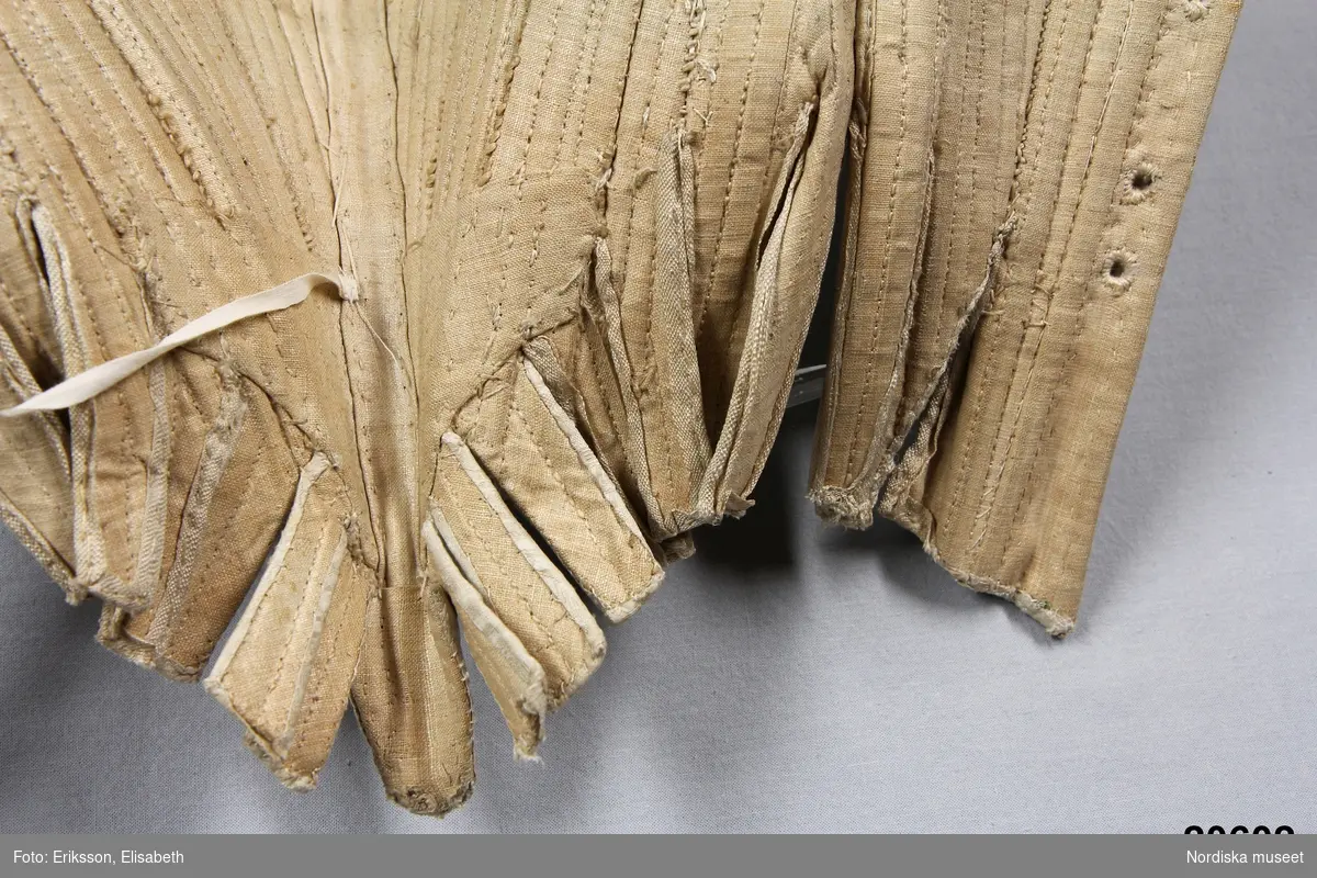 Huvudliggaren:
"Snörlif. Af hvitt siden, från 1770-talet."

Liv till hovdräkt s.k. "robe á la cour" av ursprungligen vit sidendroguet. Tyget förmodligen svensk tillverkning (Myrman?) se 1700-tals textil, A. Berchs samling, sid 171. Yttertyget är skuret i nio delar med mittstycke fram som löper ned i snibb samt fyra delar å varje sida med snörning mitt bak. Grundlivet tillverkat av oblekt linnelärft är helt styvat med spröt av valben trädda i sydda vertikala kanaler samt med en skena av järn som löper runt urringningen och planschett mitt fram. Grundlivet troligen klistrat med papper för att hålla en viss form innan yttertyget monterats på. Ärmholkar bestående av å varje sida tio hopsydda delar som lagts i veck. Tillskärningen följer helt den gängse för robe de cour. Se Waugh, Nora, The Cut of Womens Clothes, s. Längs sidorna i midjehöjd en tunn snodd avsedd att haka fast släpet i. Livet ursprungligen buret med kjol och släp i samma tyg.
/Ingrid Roos 2007-03-06