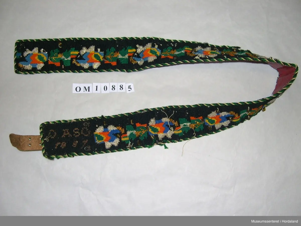 Brodert mannsbelte. Blomstermotiv med perledekor. Svart hovudfarge, blomster i grønt, gult, blått og oransje. Blanke og gullfarga småperler, nokre store, korte rørperler i gullfarge. I eine enden er det initialar og årstal, OASG 1887, sydde med perler som har rusta. Tvinna, fleirfarga snor til kanting. Raudt lêr til bakstoff. Beltet har beltespenne på baksida litt inn på beltet, og skinnreim med hol i andre enden. I tillegg er det fleire maljer og hekter.