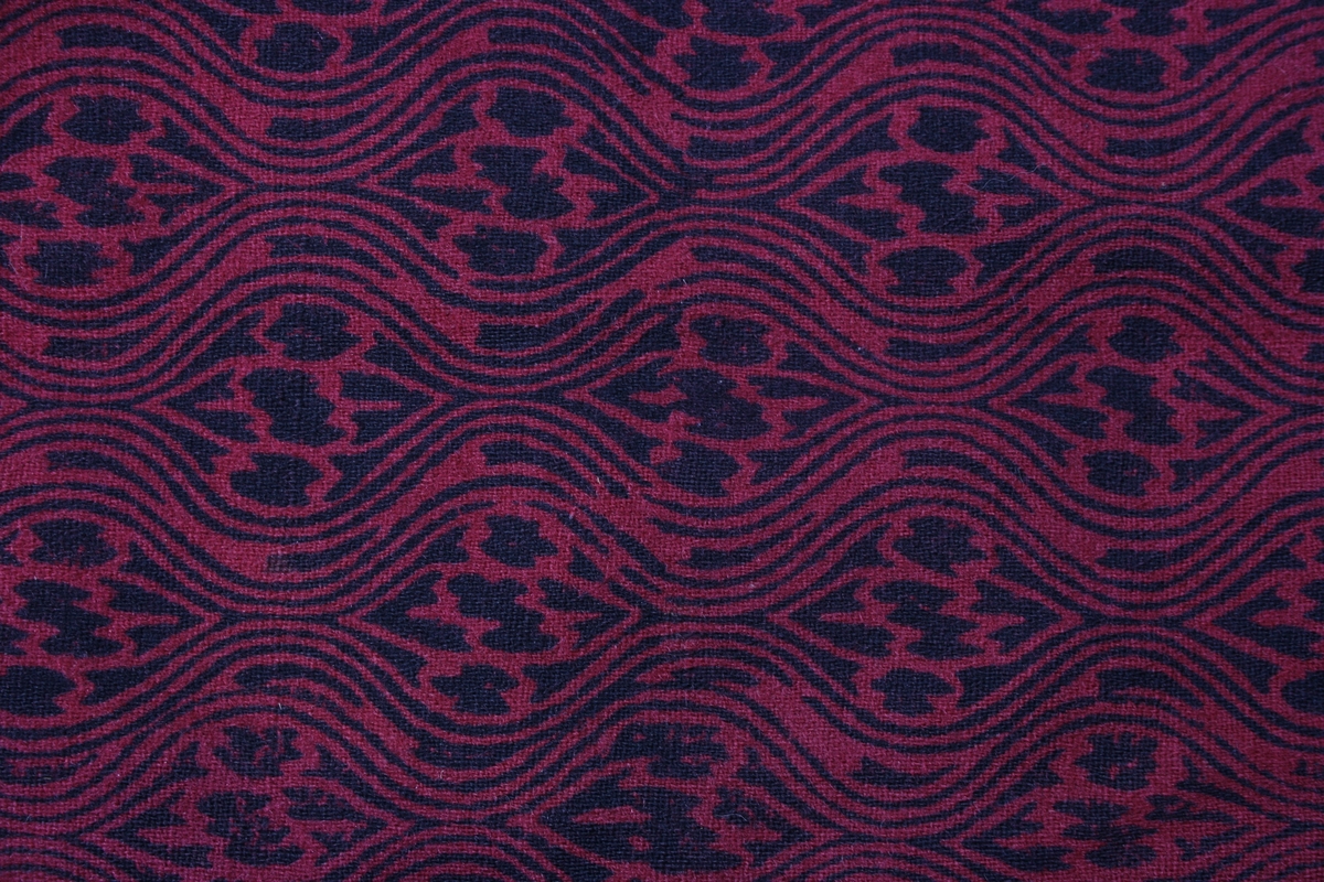 Ulveskinnspels, Lang og noe utskrådd. 
Sydd sammen av mange smale pelsstykker, to i høyden. En liten splitt midt bak. Ermer sydd sammen av mindre pelsstykker. Ermene har en liten oppbrettet fald av pelsen i nedkant, på resten av plagget er pels og fór sydd sammen kant i kant, ute belegg eller oppbrettet fald. Sjalskrage med underkrage av sort kunstpels. Underkragen har søm midt bak. 
Fóret med rødt klede med sort trykk, også i ermene.
Hele plagget er håndsydd med grov tråd, selve pelsen med ubleket lintråd. Fóret er sydd sammen, og sydd til pelsen, med ubleket eller sort bomullstråd. Fóret har sidesømmer, men ingen søm i ryggen. Ryggstykket er kvadratisk med trådrette sider. Forstykkene er trådrette midt foran og skrådd i siden. I sidene er det satt inn en liten kile nederst. 
I nedkant av kragen i venstre side er det en stor sølvhekte, gravert med blomstermotiv. Ingen tegn til annen lukning.  
Stor lærhempe i nakken.