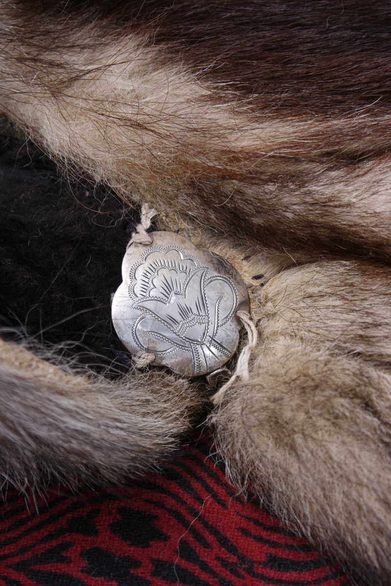 Ulveskinnspels, Lang og noe utskrådd. 
Sydd sammen av mange smale pelsstykker, to i høyden. En liten splitt midt bak. Ermer sydd sammen av mindre pelsstykker. Ermene har en liten oppbrettet fald av pelsen i nedkant, på resten av plagget er pels og fór sydd sammen kant i kant, ute belegg eller oppbrettet fald. Sjalskrage med underkrage av sort kunstpels. Underkragen har søm midt bak. 
Fóret med rødt klede med sort trykk, også i ermene.
Hele plagget er håndsydd med grov tråd, selve pelsen med ubleket lintråd. Fóret er sydd sammen, og sydd til pelsen, med ubleket eller sort bomullstråd. Fóret har sidesømmer, men ingen søm i ryggen. Ryggstykket er kvadratisk med trådrette sider. Forstykkene er trådrette midt foran og skrådd i siden. I sidene er det satt inn en liten kile nederst. 
I nedkant av kragen i venstre side er det en stor sølvhekte, gravert med blomstermotiv. Ingen tegn til annen lukning.  
Stor lærhempe i nakken.