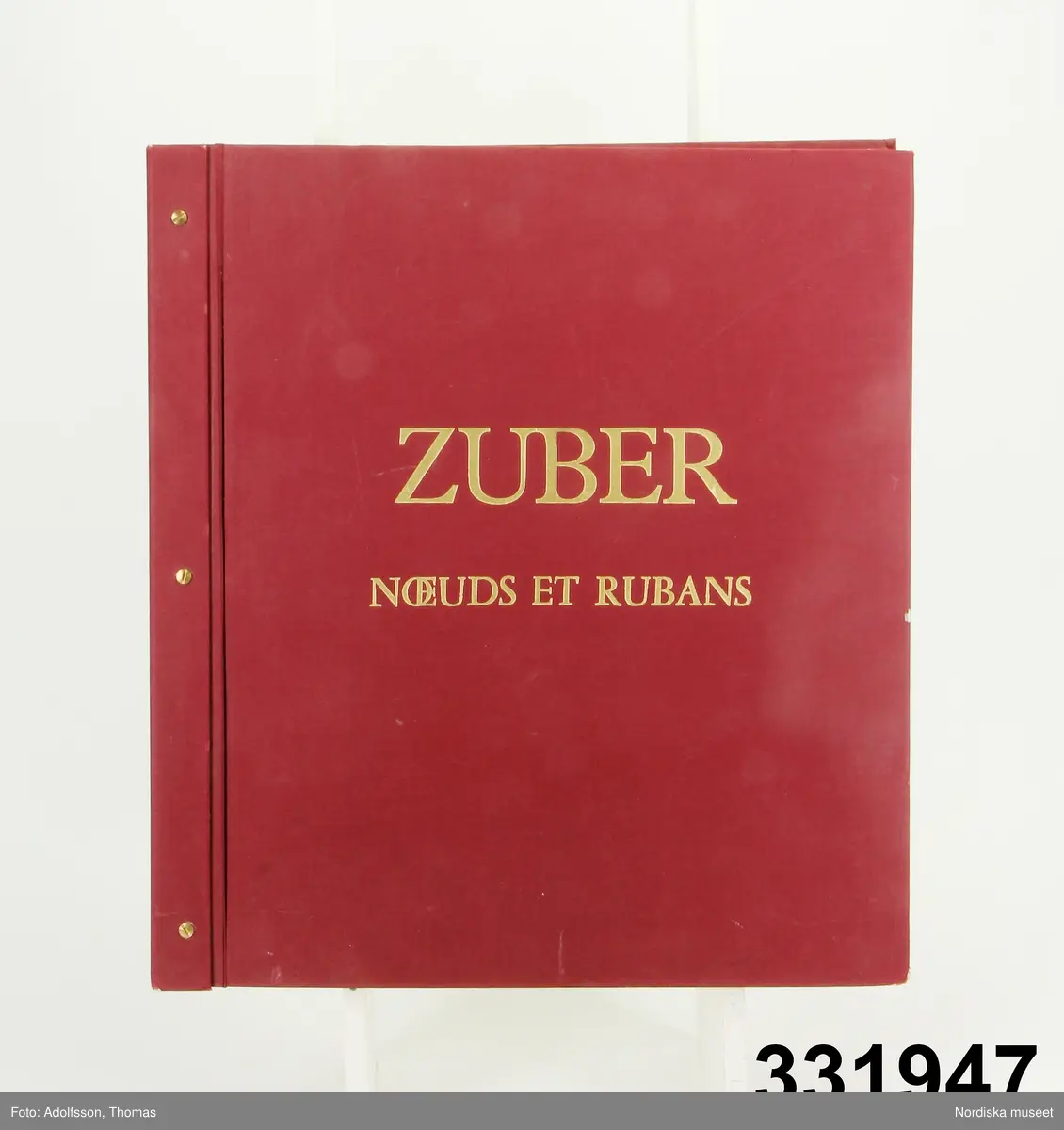 Tapetprovbok med vinröd pärm. På pärmens utsida, text i guld: "ZUBER NOEUDS ET  RUBANS" [Knutar och Band). På försättsbladet: "ZUBER & CIE Impression à la main de papiers et tissus de Haute Décoration. Rixheim-Alsace- France. Depuis 1797".

Provboken innehåller prover på handtryckta tapetbårder i olika bredder samt tapethörn, att placera där vertikala och horisontala bårder möts på väggen. Ett enda tapetmönster. Mönstren utgörs av band och rosetter, ett  mindre antal kompletterade med snörmakerier och blombuketter. Färgerna mestadels rosa, ljusblått, ljusgrönt, ljusgult.

På  pärmens insida  prislista över tapeterna i "Kollektion Noeuds et Rubans" från 1988. Priset på bårderna var 1988 196-547 kr/meter exklusive moms.Tapetens pris 725 kr/rulle.
Antalet färger avgörande för prissättningen.
/Maria Maxén 2013-05-25