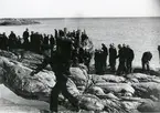 Manskap på klippstrand. Två motorbarkasser vid stranden.