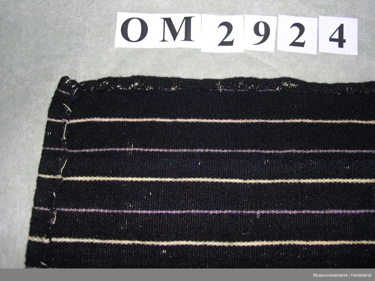 Mørkeblått forkle med kvite og lyselilla, smale og jamne striper. Linning av silkeband. To ringar med hyssing. Framme er det glatt parti og to folder på kvar side.