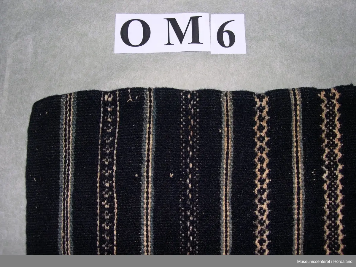 Mørkeblått, vove, stripete forkle i ull. Stripene er kvite og lyseblå. Linning med ein ring i kvar ende for feste av forkleband. Smalt, glatt stykke framme med seks folder på kvar side.