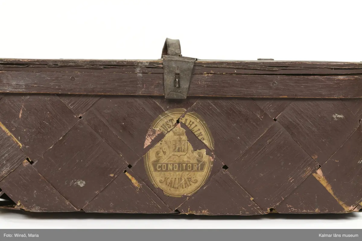 KLM 24189. Korg, konditorikorg. Fyrkantig, platt korg av flätat plattskuret spån. Korgen har ett lock med handtag och gångjärn av läder. Låsanordning av metall. Brunmålad med spår av grå färg på vissa ställen. På framsidan en tryckt etikett med text: August Sahlins Conditori Kalmar. Korgen är något trasig i botten.