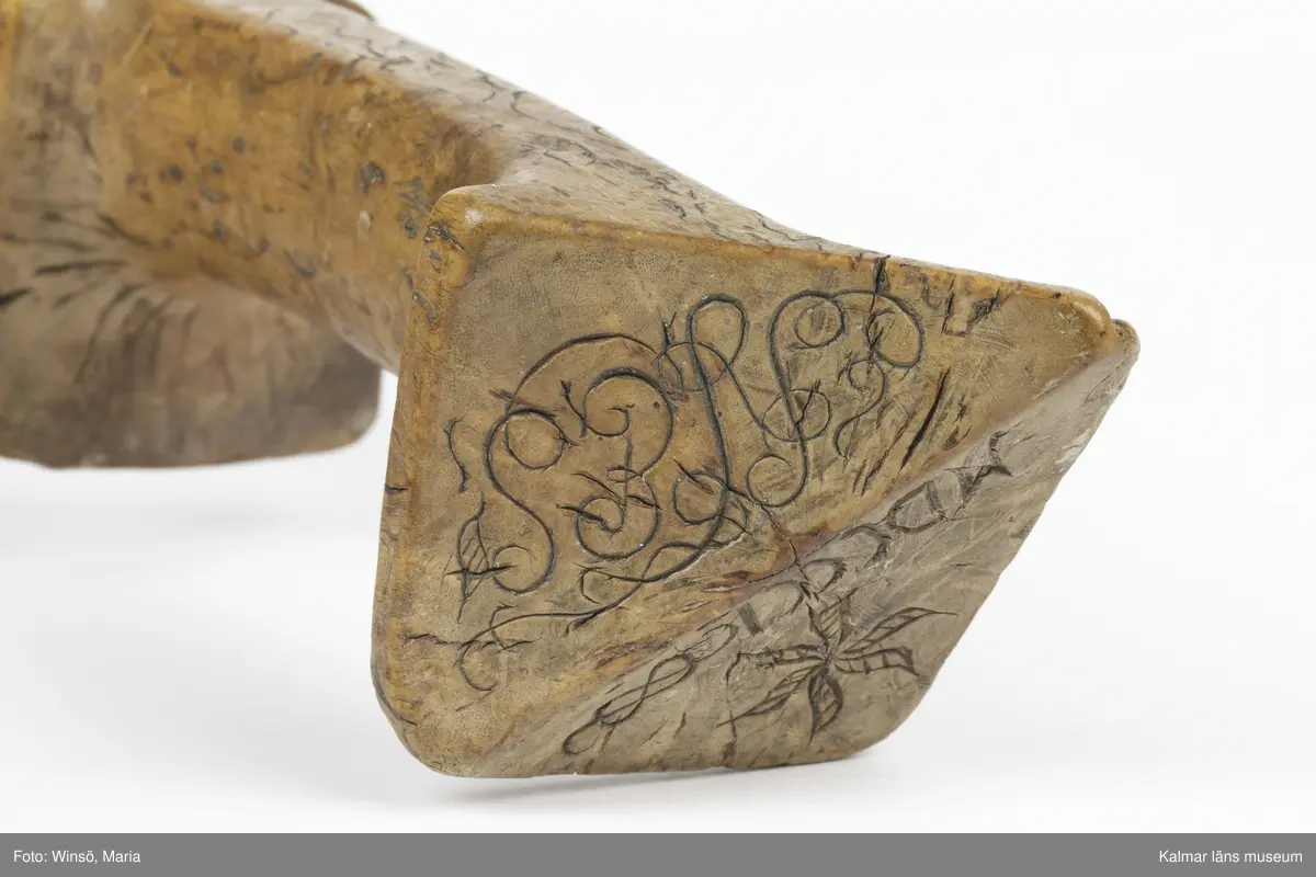 KLM 3120. Skära, vinkelskära, trä, metall. Vinkelskära med bågböjt blad av järn, fastnitat på skaftets ena sida. Skaft av masurbjörk med fyrsidig knopp med skarp mittrygg. På knoppens översida inskuret årtalet MDCCCI (1801 och blommor och rankor). På handtaget nära bladet inbränt PNS. Bladet märkt A. Användes för att skära av säden.