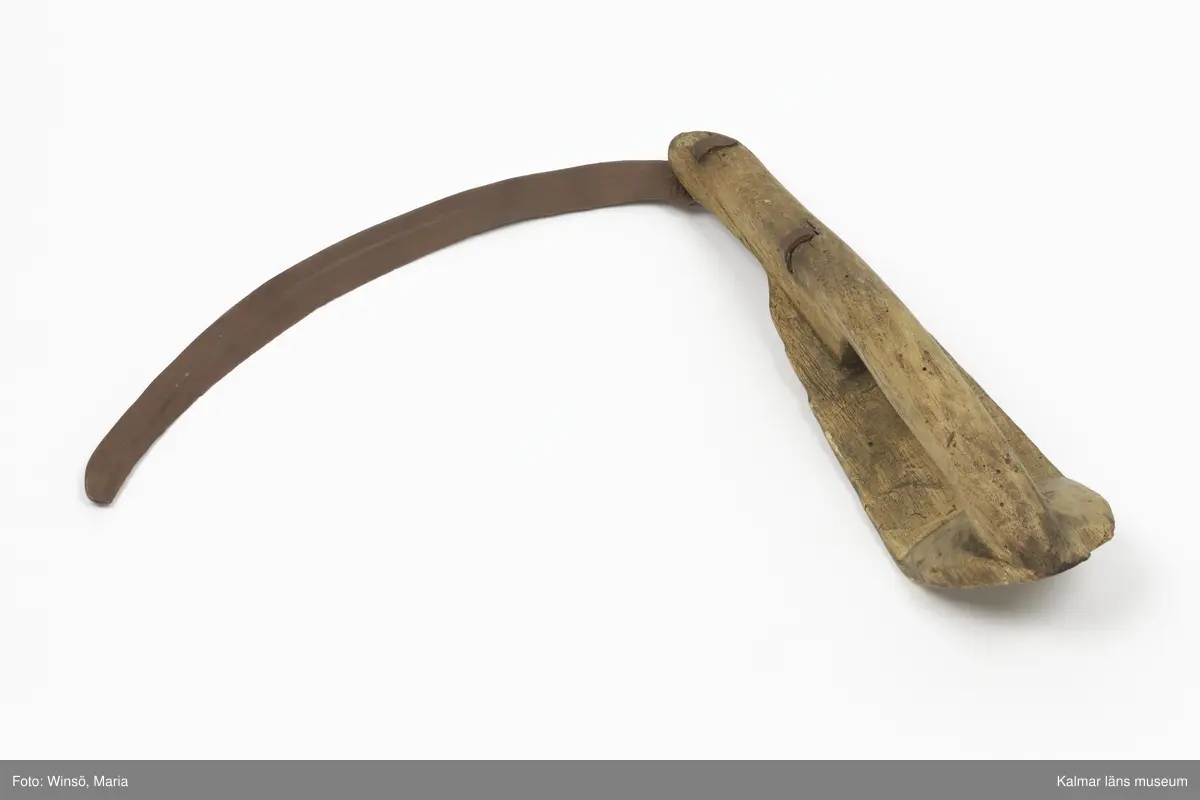 KLM 15141. Skära, vinkelskära, trä, metall. Vinkelskära med bågböjt blad av järn, fastnitat på skaftets ena sida. Skaft av trä med handbygel. Bladet är målat i rödbrun färg, för rostskydd, under senare tid. Inristning skaft: KPA, NPS HR. Stämpel blad: lis ?