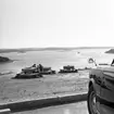 Utsiktsplatsen vid Tjörnbron. Två vuxna och ett barn sitter vid ett av borden vid vattnet. En parkerad bil syns i bildens högra kant och i bilens backspegel syns fotografen Arne Andersson.