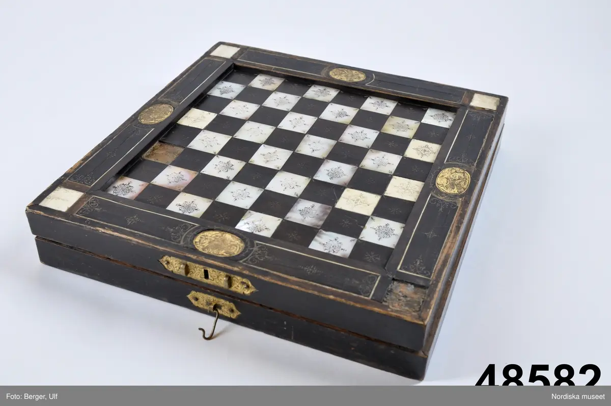 Brädspelslåda för bräde, kvarn och schack, med sju spelbrickor. Svart fanér med inläggningar av pärlemor, elfenben samt graverade förgyllda metallskivor. Blindträ av ek. (Uppgifter från katalogkort). Tillhörande träask för förvaring av spelpjäserna.
