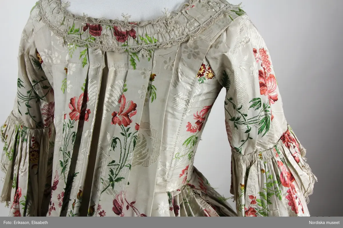 Äldre katalogkort: 
"Robe och kjol  av vit sidenbrokad med blommor i rött, gult m.m. Robe med dubbla watteauveck och engageanter av vita knypplade silkespetsar i mångdubbla rader. Livet fodrat med vitt linne med fiskben i sömmarna. Bröstlapp saknas.
Tygets vådbredd 50,5 cm. Tygåtgång ca 13,5 meter."

Bandgarnering av veckad remsa av tyget kantat med uddganser i vitt silke.
I utmärkt fint skick.
/Berit Eldvik 2009-01-09