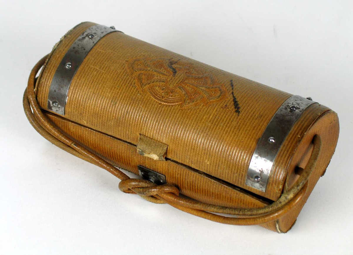 Halvsylinderformet syskrin i brunt skinn. Dobbel skinnsnor festet i kortendene. Lokket har metallbeslag og preget dekor. Syskrinet inneholder nålpute, pren, saks og foldekniv.