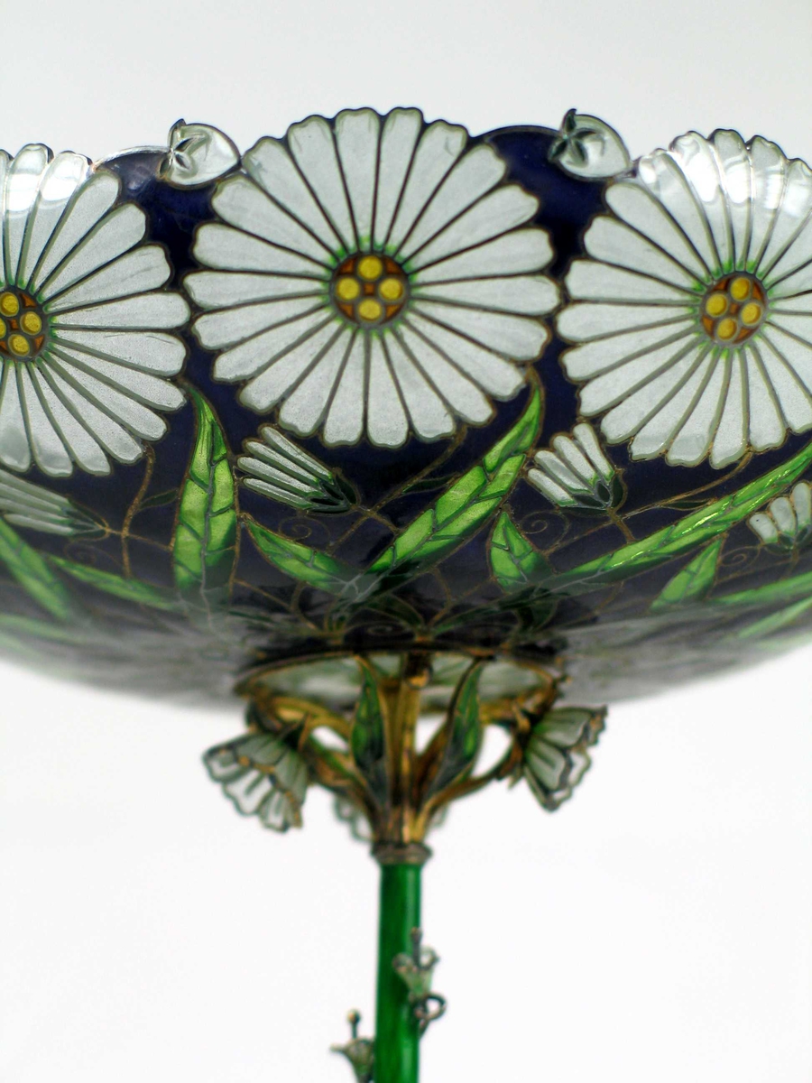Skålformet vase med lang, tynn stett og opphøyet fotstykke. Foten er laget i sølv og grønn emalje. Fra denne slynger det seg blomster, knopper og bladverk oppover stetten. Oppunder den flate glasskålen er det tre blomster, knopper og blad. I selve skålen er det innerst en rosett med gullkule i midten. Videre er det samme mønsteret gjentatt på mørk blå bunn; to grønne blad, en knopp og øverst en utsprungen margeritt.
