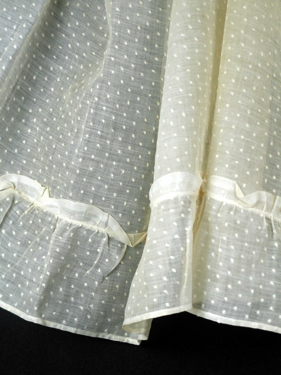 Hvit kappe i bomull med innvevde prikker. Smal maskinsydd fald på tre sider. Nederst påsydd 12 cm bred rynkekappe der rynkehode har jarekant.