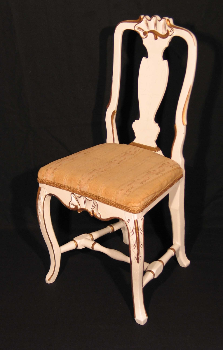 Hvitmalt stol i bjørk med utskåret muslingmønster på toppstykke og sarg. Også utskåret ryggbrett og sprosser. Svungne forben. Bronsemalte detaljer. Setet er trukket om i brokadestoff (skaffet av Dagny.)