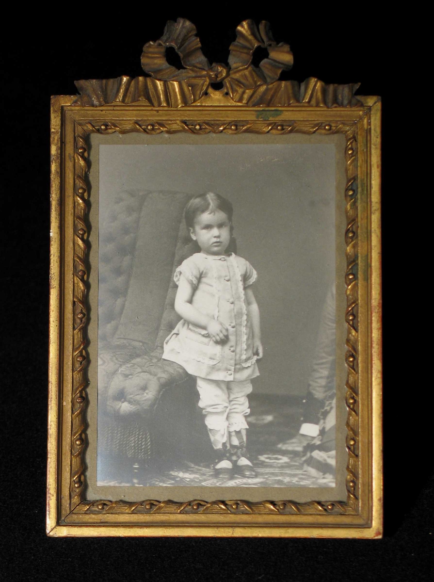 Jente fotografert i atelier. Står lent mot en stoppet stol. Bildet er beskjært; en arm til høyre.