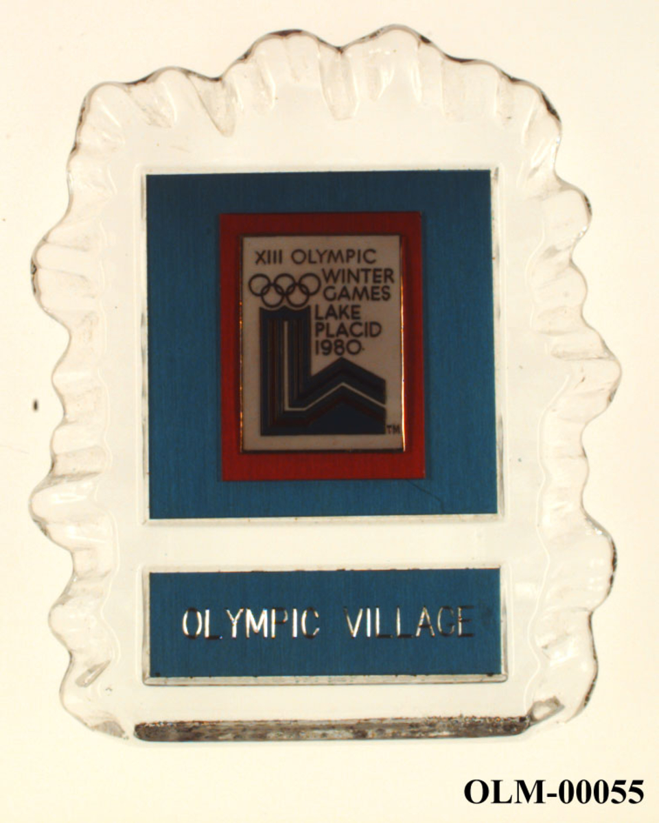 Brevpresse av glass påfestet en plakett av metall. Plaketten har emblem for de olympiske vinterleker i Lake Placid i 1980.