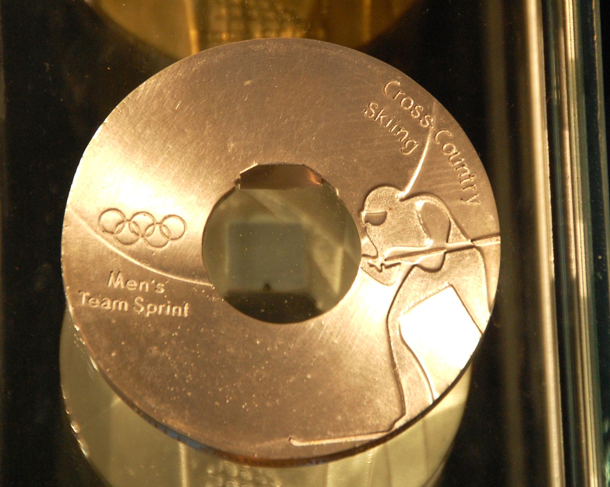 Sølvfarget medalje med motiv av en langrennsløper og de olympiske ringene. 