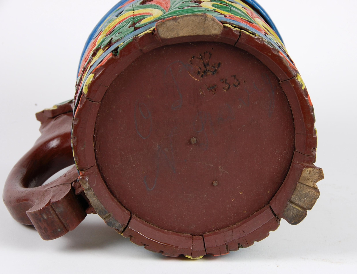 Kanne av staver. Lokk med gule, grønne og røde blader, blåmalt kant. Korpus med en blåmalt metallgjord oppe og nede, gule, grønne og røde akantusblader, brunmalt hjerteformet felt med innskriften 1865. Brunmalt hank. Tre røde og gule føtter.