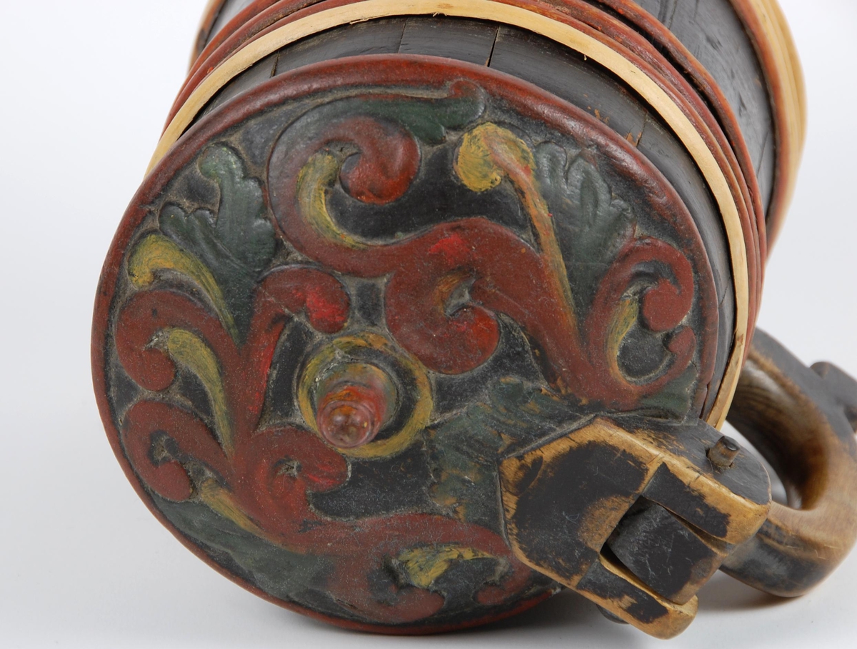 Kanne av trestaver. Lokk med rødt, gult og grønt akantusskurd. Korpus med fire gjorder oppe og nede, fire av gjordene er av nyere dato, sotmalet. hanken er skåret ut av en stav.
