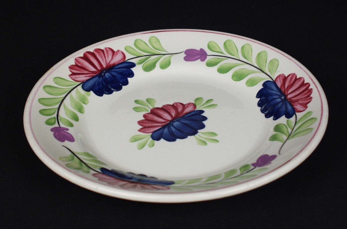 Deler av servise i steintøy og flintgods. Godset er hvitt med malt blomsterdekor. Rød og blå blomst i midten med en ranke av grønne blad.