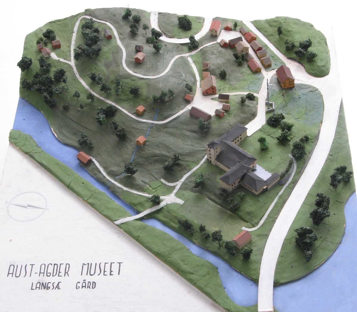 Plan og modell over Aust Agder Museet,  1952.   Furukasse  med   finerlokk,  I kassen en modell av Langsæterrenget,  med Barbuelva. Hovedbygningen fra 1858 er borte,  museumsbygget er større enn nå. Rundt i  terrenget spredte tun og bygate = Aust  Agder By og Folkemuseum (som omtalt bl. a.  i hefte 1934, Albert Uglands plan for museet).  