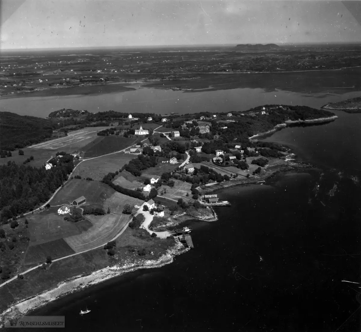 Aukrasanden og Holmesundet og utover mot Varhaugvik og Nerbø, med Bud i bakgrunnen. Midt på bildet Aura Kirke og Aukra Fiskarfagskole. I forkant ein fiskebåt og ferga som ligg ved kai.