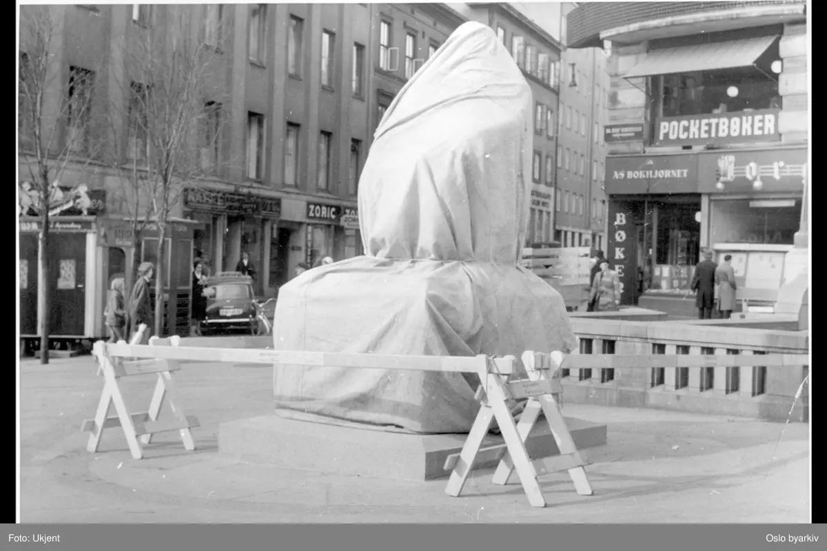 Bronsestatuen av Christian Krohg på Stortings plass innpakket i lerret og avsperret innen avduking. Arbeidergata med diverse butikker i bakgrunnen. Reklameskilt. Bokhandel (A/S Bokhjørnet) og utstillingsmontre til Morgenbladet bak til høyre i Centralgården.