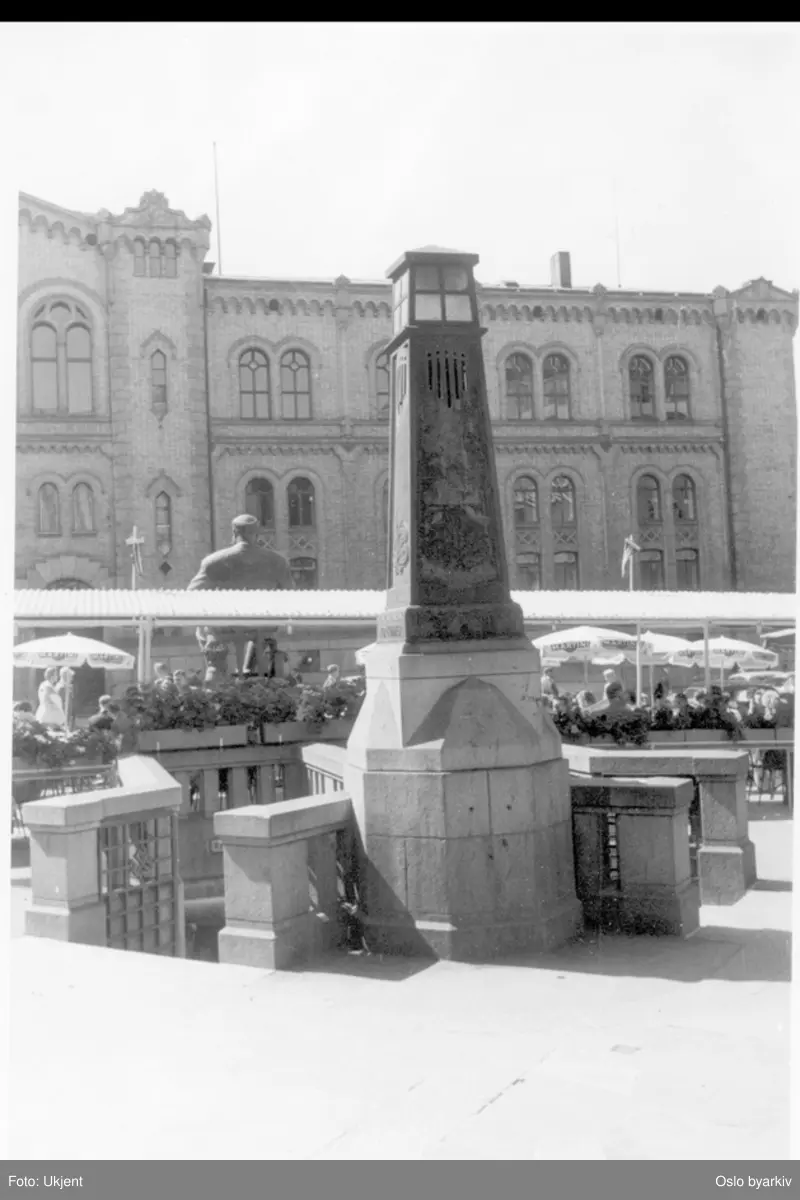 Nedgang til toaletter ved lyktetårn på Stortings plass. Uteservering ("Dasslokket") under parasoller.Stortingsbygningen langs Karl Johans gate i bakgrunnen.