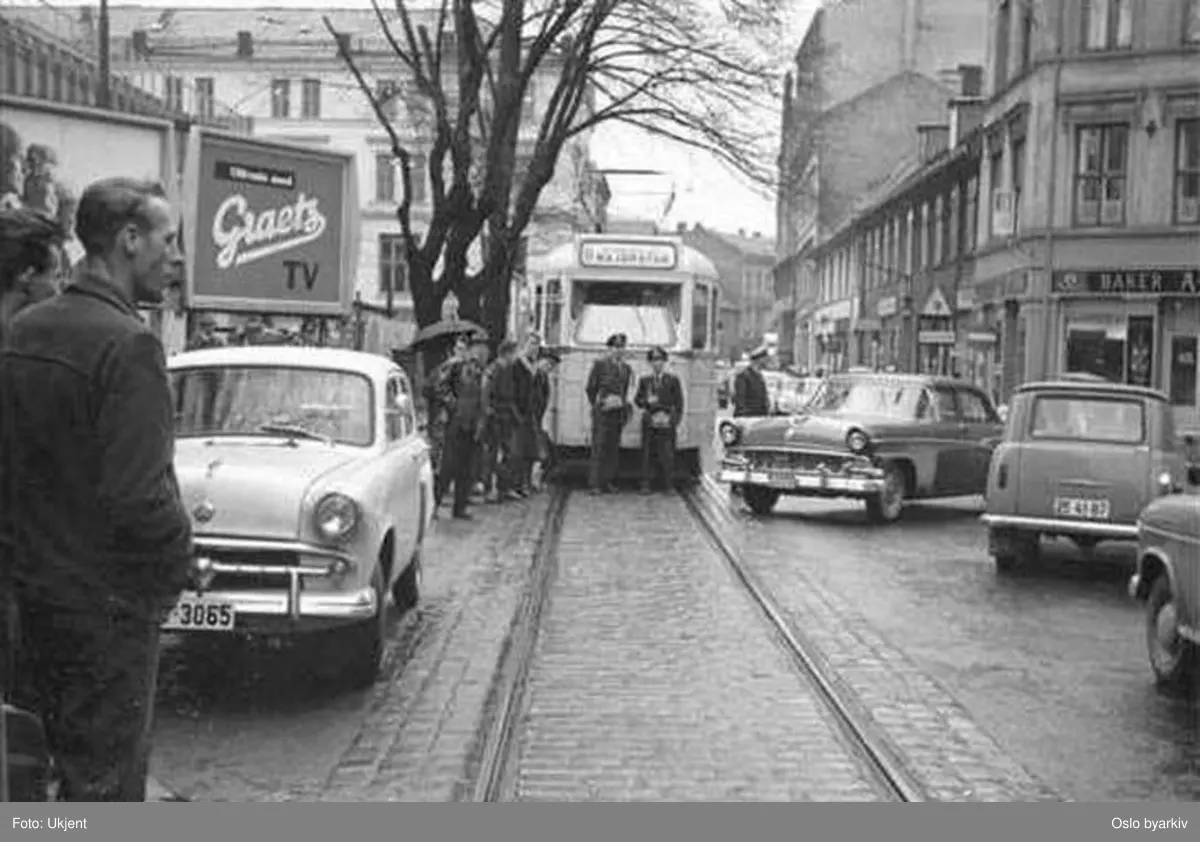 Oslo Sporveier. Trikk motorvogn, trolig 211 type Høka MBO, linje 11 i Pilestredet. Feilparkert Moskovitch hindrer videre kjøring. Betjeningen står utenfor og venter på hjelp til å flytte bilen. Tilskuere, biler, TV-reklameplakat.