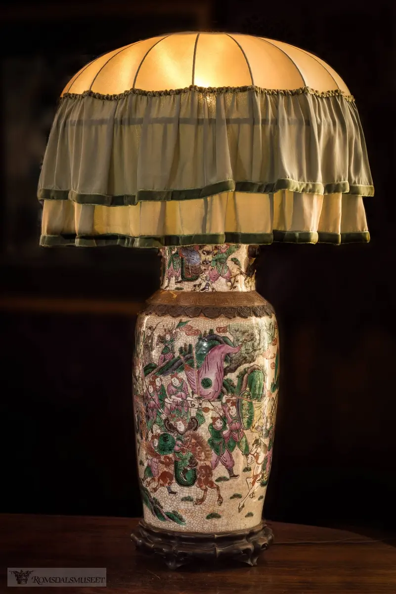 Chateauet..Bordlampe på flygelet. Lampa er en kinesisk porselensurne.som står på en liten tresokkel. Skjermen er som seg hør og bør av silke, et lys grønt stoff med legg og rysjer. Kinesisk porselen fra de gamle keiserdynastiene har i flere 100 år vært samleobjekter, og har gitt status i borgerlige, vestlige miljøer..Dette er nok ingen verdifull, gammel urne, men en nylaget innredningsartikkel fra tida Chateauet var ferdig og skulle innredes. Sannsynligvis produsert i Kina. Den var garantert en kostbar investering, og ga interiøret et preg av tung eleganse og klasse..Kineserier var på moten på denne tida. Etter dagens trender er lampa neppe særlig vakker, men desto mer tidstypisk. .(Se Romsdalsmuseets årbok 2012)