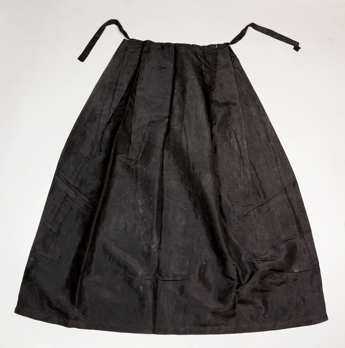 Sort forkle i tynt, sort stoff, med sløyfebånd til knyting, som er en forlengelse av øvre kant på forkleet. 