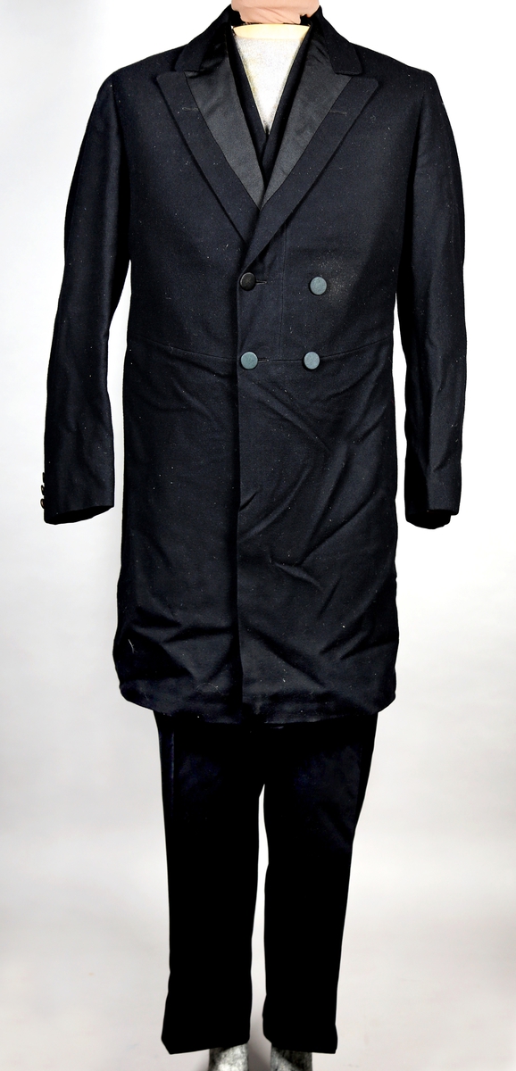 Svart dress med vest og lang jakke. Jakka er dobbeltspennet og kneppes med fire knapper, som er trukket. Jakka har splitt bak. Den har silkefôr, bortsett fra i ermene. Buksa har både plastknapper og metallhekte, og tre lommer. 