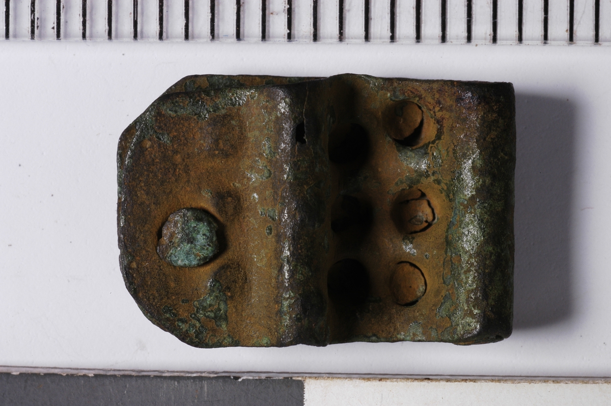 MALV: L. 15 MM H. 10 MM, BESTÅR AV BØYD, REKTANGULÆR BRONSEPLATE, AVRUNDET IENDENE. NAGLEHULL MED NAGLE. 6 FORDYPNINGER ER HAMRET INN, ANTAGELIGSOM ET EKSTRA FESTE I REIMA. 6 MINDRE DITTO KAN OGSÅ ANES. STOLPEN ISPENNA SITTER FORTSATT I. 