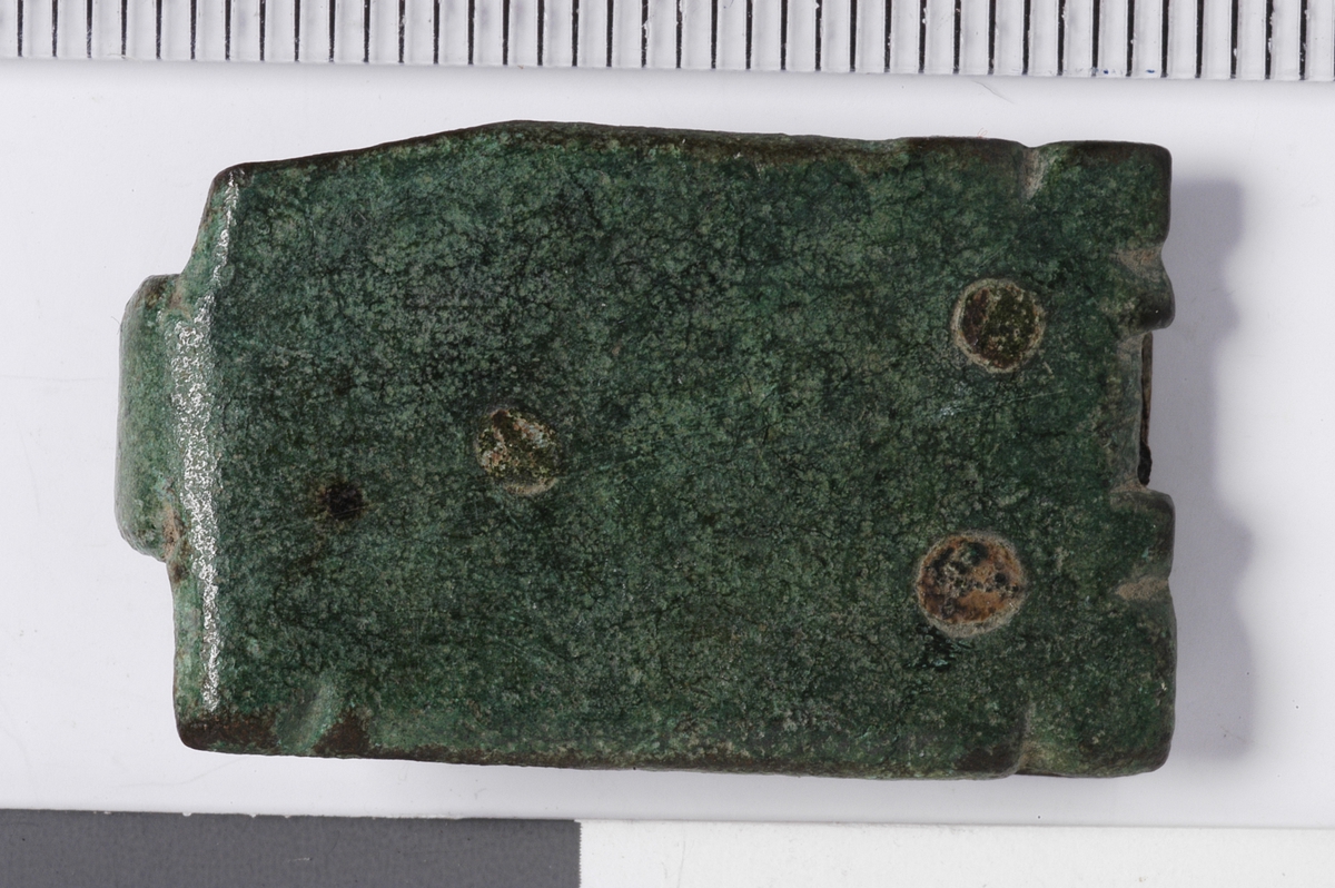 MALV: L. 28, 2  B. 18 MM, BANDFORMET MED HEMPE I ENE ENDEN. PÅ BAKSIDEN PÅSATTEN TYNN PLATE MED NAGLER. RESTER AV PERMEN SITTER IGJEN MELLOM SPENNEOG PLATEN, UTEN DEKOR. 