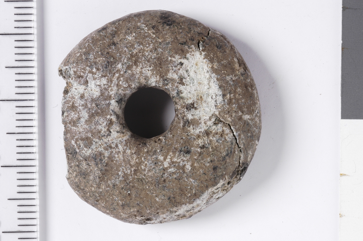 Sirkulært spinnehjul i stein, med en skade i kanten. Gjenstanden har et tilnærmet halvkuleformet utseende med flat underside og buet overside.