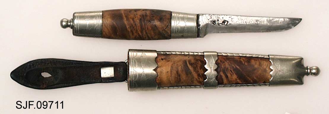 Tollekniv med slire.  Slira er signer "T. B. ", som skal være initialene til Trygve Brenna fra Åmot på Modum.  Bladet er 8, 2 centimeter langt og 1, 9 centimeter bredt ved brystinga.  På den bakre delen av nakken, som er om lag 0, 5 centimeter bred, er det filt to tverrstreker og et kryss.  Høyre bladside har tre innstemplete initialer, antakelig "Ø. L. S. ", innskrevet i ei buet ramme.  Denne signaturen ble brukt av eggsmeden Knut Knutsen Øyo (1860-1921), og seinere av firmaet til sønnene Lars, Thomas, Knut og Olav Øyo, «Øyo ljåsmie».  Dette navnet ble brukt etter 1. verdenskrig.  Firmanavnet ble for øvrig endret til «Brødrene Øyo» i 1941, som en konsekvens av at produktspekteret bedriften kunne levere var blitt bredere, og av at ljåblader ikke lenger var det dominerende produktet.  Bladet på denne kniven bærer preg av å være slipt mange ganger. 

Skaftet er lagd av flammebjørk Det er snaut 9, 5 centimeter langt og har en noe konveks form i lengderetningen og et ovalt tverrsnitt (snaut 2, 5 X 1, 95 centimeter på midten).  Begge ytterender er forsterket med 2, 0 centimeter brede nysølvholker med inngraverte linje-, bue- og rombemønstre.  I enden av bakholken er det påsatt ei rombeformet plate, som den dreide knoppen enden av knivbladets tange er forankret i er festet mot.  Tredelen av skaftet har en lys gulbrun farge som muligens skyldes forsiktig beising.  Kniven - inkludert blad, skaft og knopp - er 18, 7 centimeter lang.  

Kniven har treslire, lagd av flammebjørk og bundet sammen av nysølvbeslag.  Slirebeslaget består av to spenger langs sidene, som holdes ihop av tre holker: en munningsholk (øverholken), en midtholk og en doppsko.  Munningsholken har samme dekor som holkene på skaftet, altså linjemønster (parallelt med overkant), bue- og rombemønster (skravert).  Nederkanten på denne holken har et profilert mønster på den sida som skulle vendes fram når kniven ble båret i bukselinninga.  På baksida er det påloddetei hengsle for en beltestropp med "smikk" av brunsvart lær.  Den bakre delen av denne stroppen har spalte for på- og avknepping, den fremre et hull for forankring av kniven i knoppen i skaftenden.  Midtholken på slira har inngravert to "skraverte" romber, og har profilerte kanter på forsida av slira.  Baksida er glatt, bortsett fra at det er her knivmakeren har gravert inn initialene sine med ei kort pil på hver side.  Doppskolen er tilnærmet rektangulær og har en trefliket "rombe-rosett" som en profilert overkant som dekor på forsida.  I enden av doppskoen er det påsatt en dreid knopp av samme type som den som er brukt i øverenden av skaftet. 

Redskapet veier 135 gram, herav sjølve kniven 70 gram. 