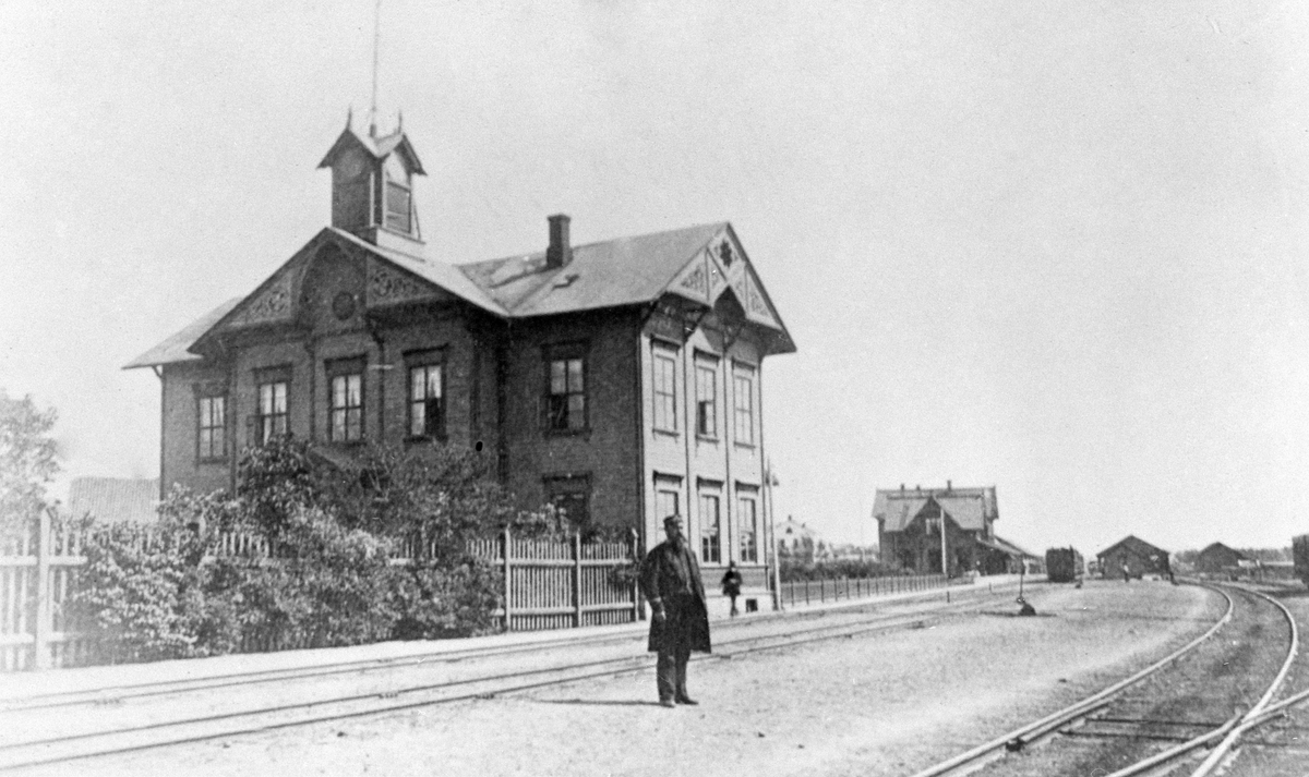 Hamar jernbanestasjon, Hamars første stasjonsbygning fra 1862, åpnet 23. juni, Grundsetbanen Hamar- Elverum, i bakgrunnen sees Hamars andre stasjonsbygning som ble tatt i bruk i 1880 for Hamar-Eidsvoldbanen, i forgrunnen ingeniør/stasjonsmester Christian Fredrik Lund (1838 -1886), (gift med Anne Christine f. Amundsen)