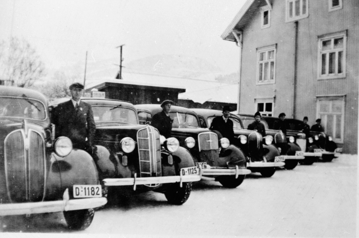 7 drosjer med sjåfører oppstillt.  Fra venstre er Martin Sørum med D-1182 Plymouth 1937, D-1125 Dodge 1936, Johan Lindbak med Chevrolet 1936, Olaf Stigen D-2565 Plymouth 1938, Guttorm Lehne D-4114 Plymouth 1937, Johannes Røseth D-2145 Dodge 1938, Johannes Lillesveen D-4414 Plymouth 1937, Mikael Bakkerud ved dosjeholdeplassen foran Sørumgården, Furnesvegen, Brumunddal.