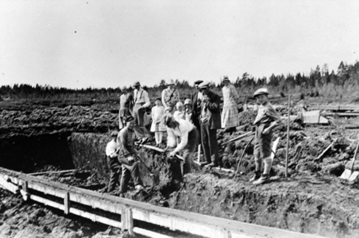 Voksne og barn arbeider på Leikmyra torvfabrikk. Brenntorvgruve i Furnes Almenning. Syverin Myhre, Egil Mikkelsen, Kåre Godager. Tilskuere er garver O. Kirkeby med familie, Petter Kirkeby med pipe og Erika Sveum. Gutten til høyre er Kristian Sveum.
